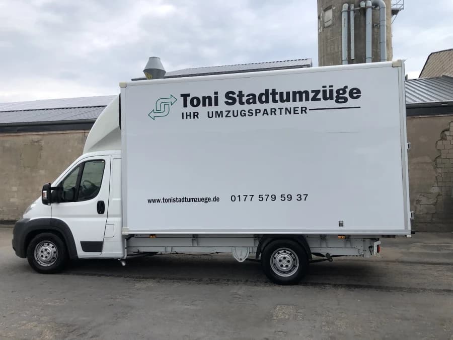 Weißer Kastenwagen mit Ladebordwand von der Seite. Auf dem Koffer stehen der Schriftzug „Toni Stadtumzüge – Ihr Umzugspartner“, die Internetadresse und die Telefonnummer des Betriebs.